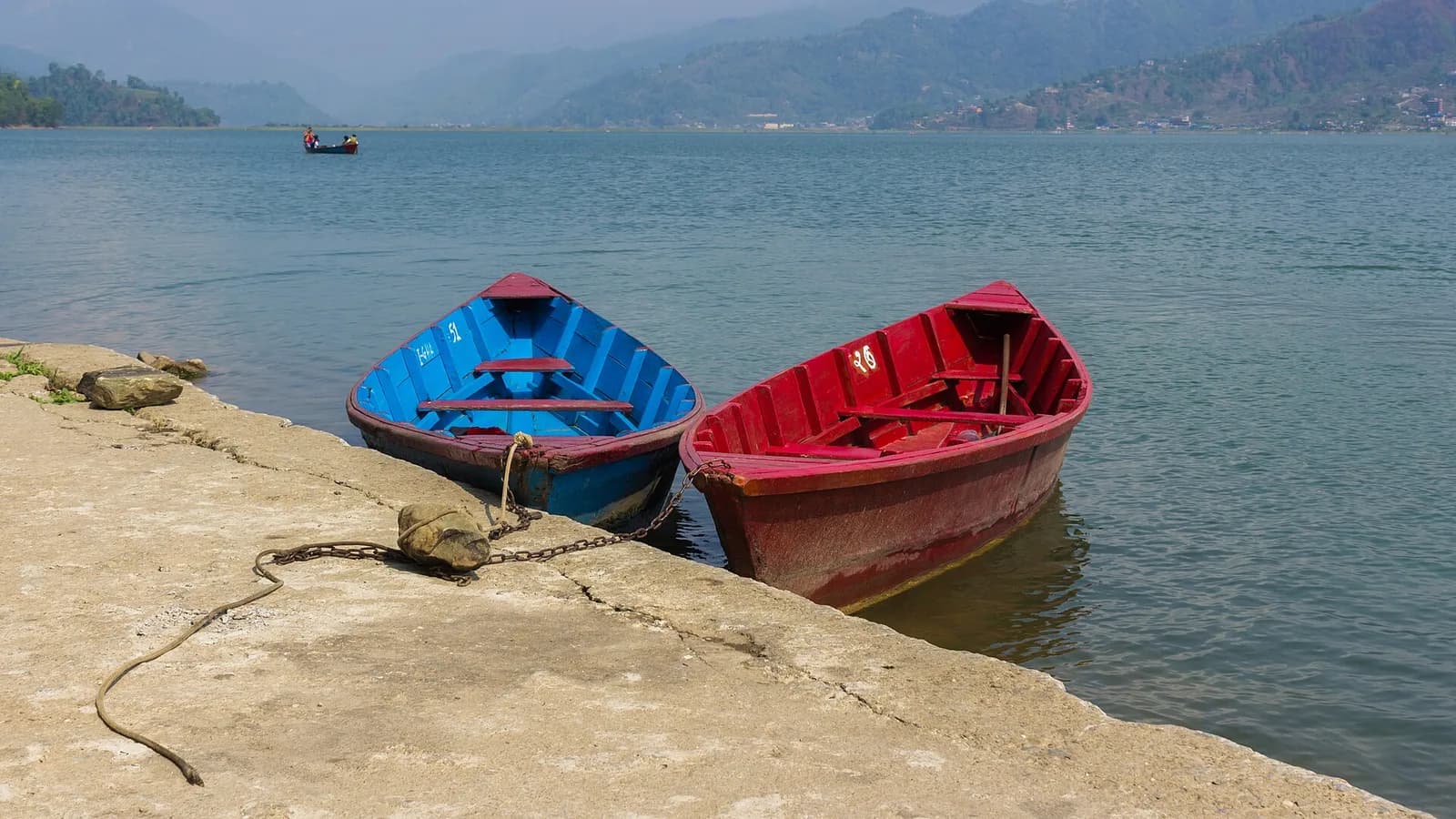 Phewa Lake in Pokhara, Nepal, with calm green water, wooden boats near the shore, and forested hills rising behind.