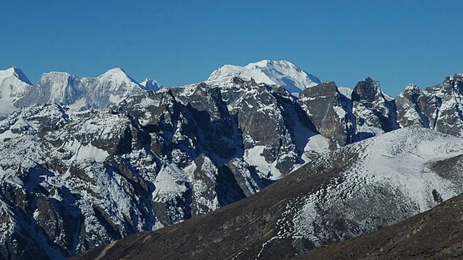 A wide view of jagged, snow-covered Himalayan peaks rising in a continuous wall under a clear blue sky