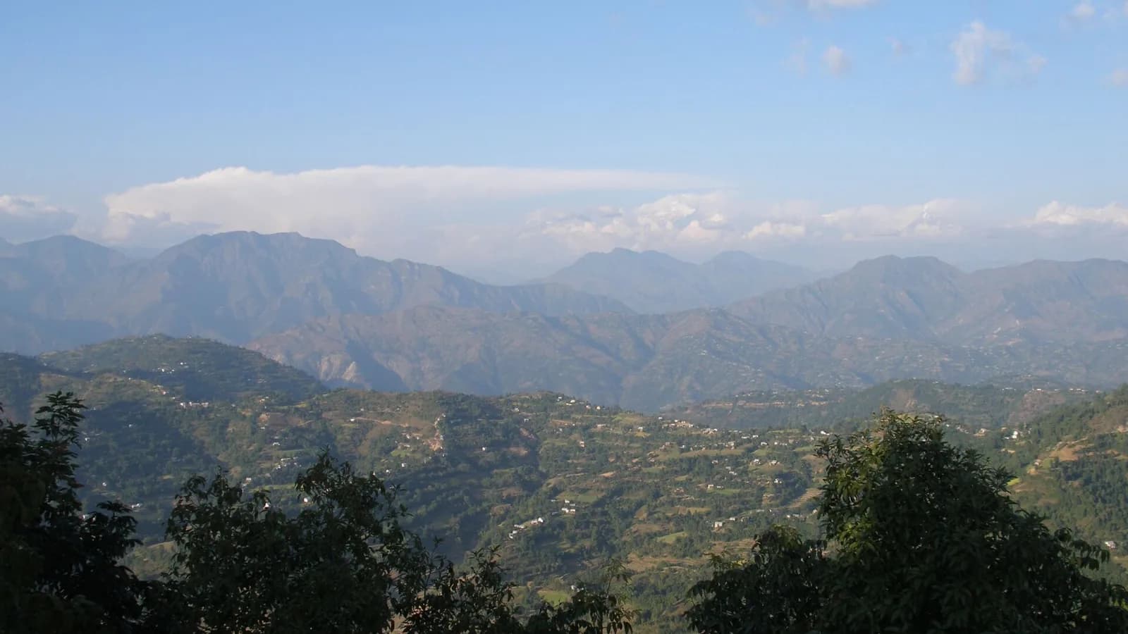 A Nepali Himalayan landscape with forested foothills and distant snow-capped peaks under cloud, similar to the high country around Kalinchok